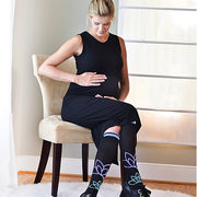 Seated woman holding pregnant belly wearing black compression socks with lotus flowers and light blue & purple accents